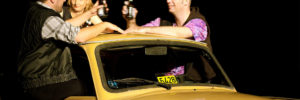 Drei Darsteller sitzen auf den Türen eines Taxis (Trabant) und prosten sich mit Bierflaschen zu