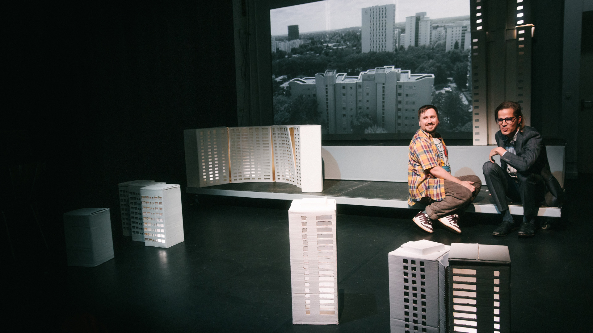 Zwei Personen sitzen auf einer Bühne zwischen weißen Architekturmodellen, im Hintergrund ist eine Stadtansicht zu sehen.
